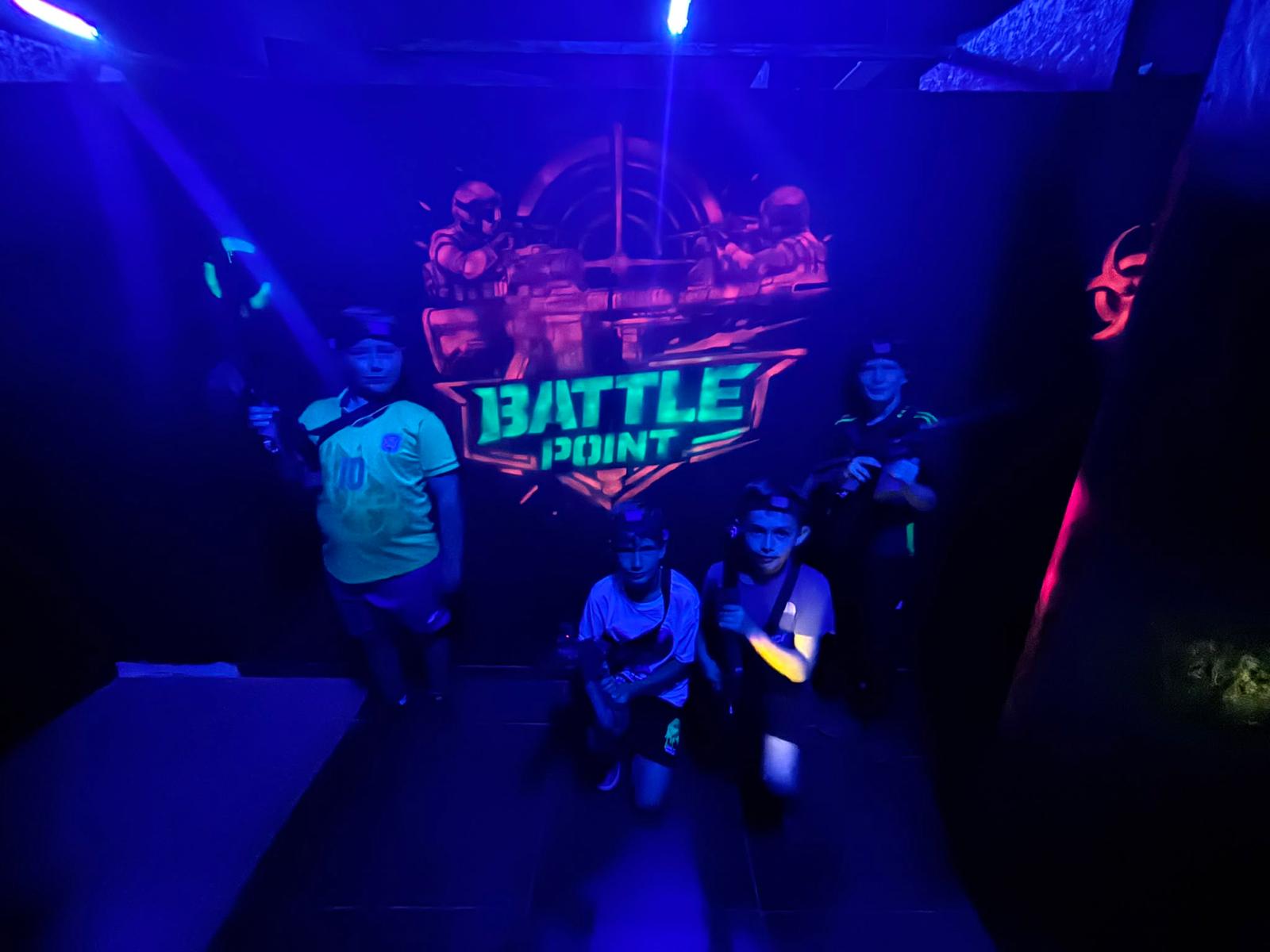 Grupa dzieci pozuje z karabinami laser tag na tle muralu Battle Point w niebieskim świetle UV